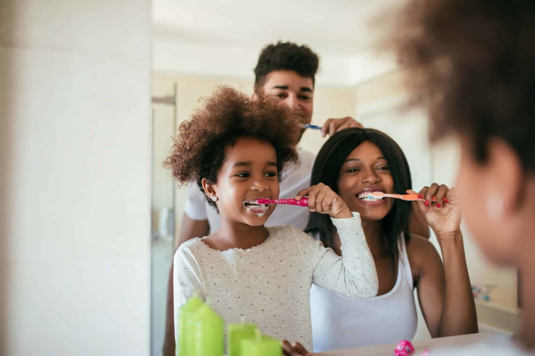 A Family Of Three Standing In A Bathroom, Brushing Their Teeth Together.